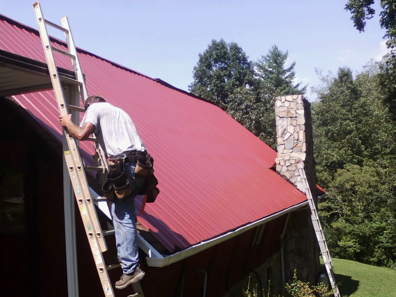 Roofer on a ladder beside a red metal roof and stone chimney — Soffit & Fascia Repair by Roofing Brothers in Oakesdale